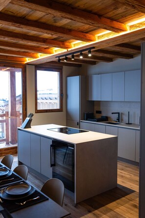 A modern kitchen with a central island, wooden ceiling beams, and a view through a window.