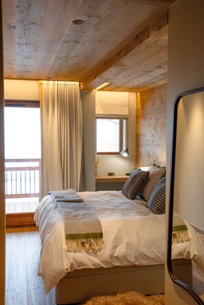 A bedroom with a wooden ceiling, a bed with a white duvet, a window with curtains, a bedside table with a lamp, and a mirror on the wall.