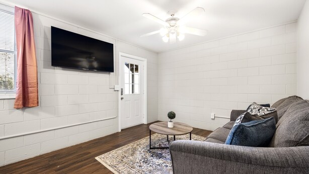 Un salon avec un canapé gris, un téléviseur à écran plat fixé au mur, un ventilateur de plafond et un tapis à motifs.