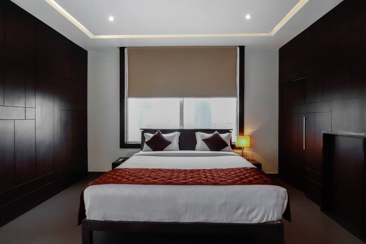 A modern hotel room with a large bed, dark wood paneling, a large window, and a nightstand with a lamp.