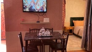 Habitación de hotel con una mesa de comedor, sillas, televisión montada en la pared, cama con cabecera y mesita de noche.