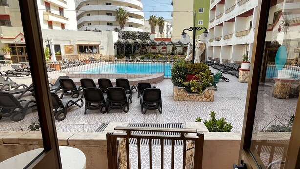 A view of a hotel courtyard with a swimming pool, lounge chairs, and a restaurant. - Cozy poolside apartment with 5 beds (San Pawl il-Baħar)