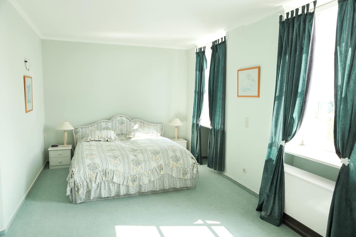 A bedroom with a bed, two bedside tables, a window with curtains, and a wall-mounted light fixture.