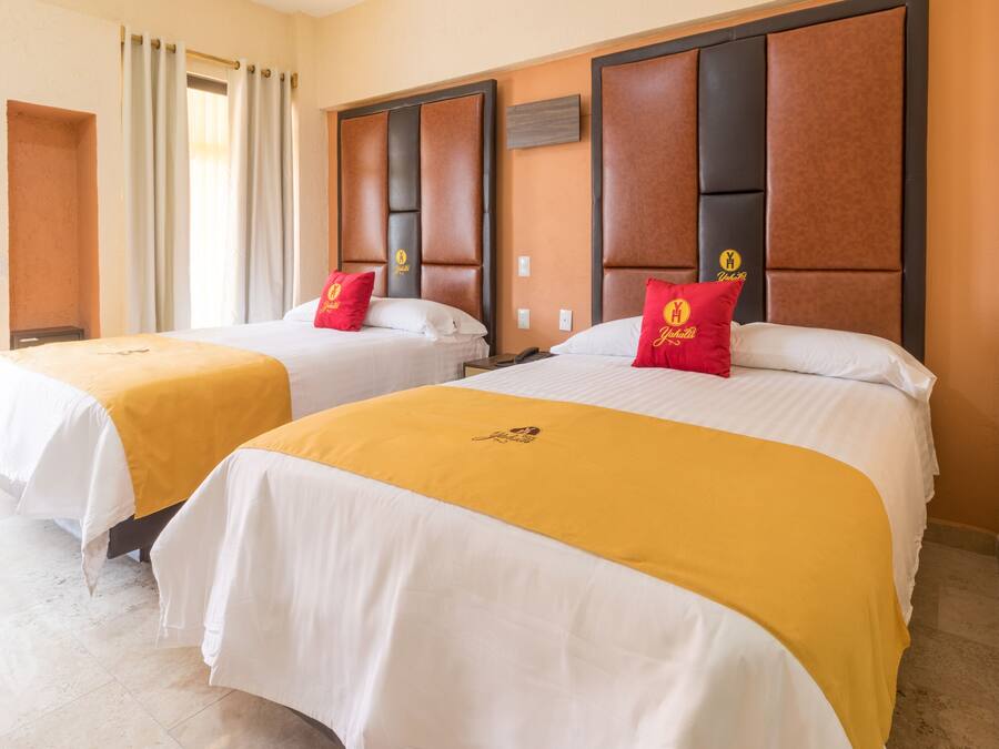 A hotel room with two beds, each with a yellow bedspread and a red pillow. The room has a wooden headboard and a small table between the beds.