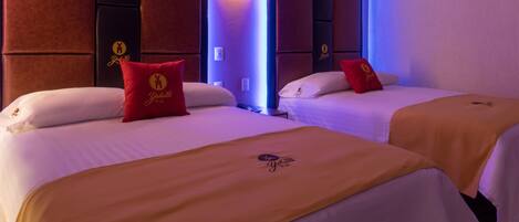 A hotel room with two beds, each with a red pillow and a gold blanket featuring the hotel's logo.