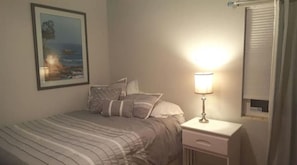 A bedroom with a bed, pillows, a bedside table with a lamp, a framed picture on the wall, and a window with blinds.