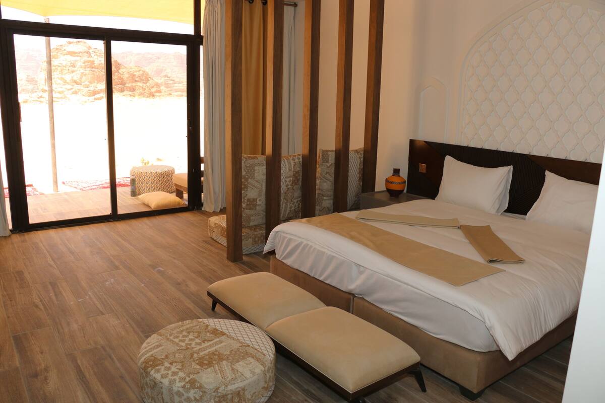 A hotel room with a large bed, a bench, and a view through a sliding door to an outdoor area.