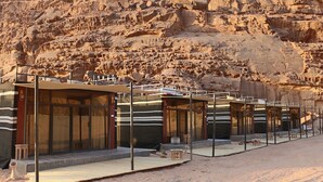 A row of modern, glass-walled rooms with metal railings, set against a backdrop of rugged, rocky cliffs. - Rum Imagination Luxury Camp (Wadi Rum)