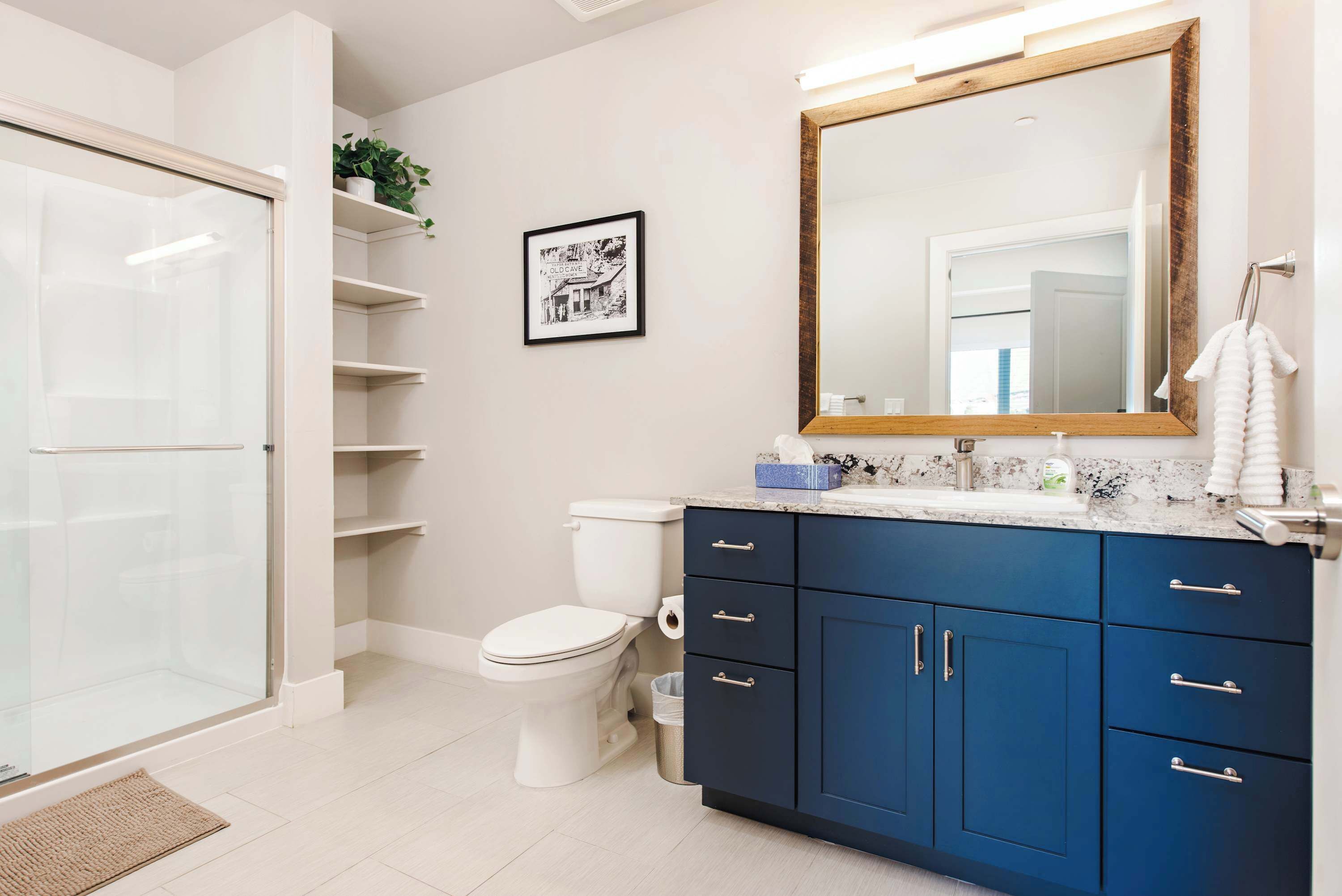 A modern bathroom with a glass shower enclosure, a white toilet, a blue vanity with drawers, and a large mirror.