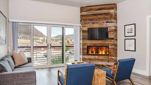 A living room with a fireplace, a sofa, two armchairs, a TV mounted on a wooden wall, and a balcony with a view of the mountains.