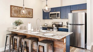 A modern kitchen with dark blue cabinets, stainless steel appliances, a marble island with bar stools, and pendant lighting.