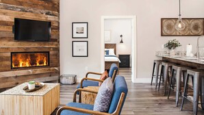 A modern living room with a fireplace, a TV mounted on a wooden wall, a seating area with a blue sofa, and a kitchen area with bar stools.