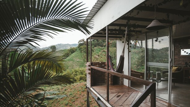 A wooden deck with a view of a lush landscape, a glass door leading to an interior space with a brick wall and a hanging light fixture. - San PEREGRINO (Manizales)