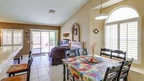 A dining area with a table covered in a colorful quilt, surrounded by wooden chairs. There is a bench and a sofa in the background, and a window with blinds on the right.