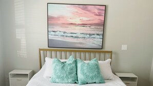 A bedroom with a bed, a headboard, two bedside tables, and a wall-mounted painting of a beach scene. - Bikes, Beaches and Quaint Floridian Downtowns (Palm Harbor)