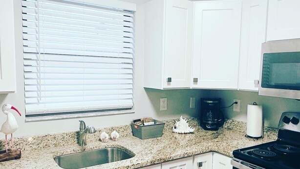 A kitchen with white cabinets, a granite countertop, a sink, a stove, a microwave, and a window with blinds. - Bikes, Beaches and Quaint Floridian Downtowns (Palm Harbor)