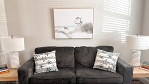 A dark grey sofa with two patterned pillows, flanked by two white lamps with textured bases, against a wall with a framed picture of a beach scene. - Bikes, Beaches and Quaint Floridian Downtowns (Palm Harbor)