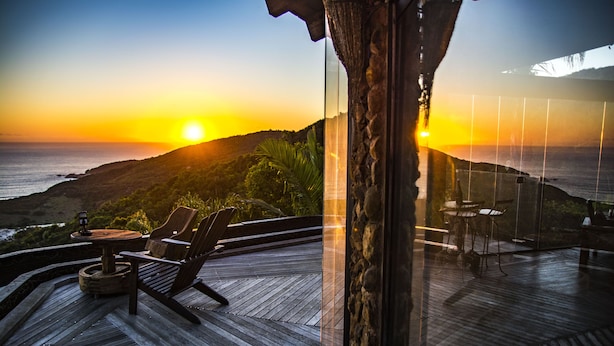 A wooden deck with two lounge chairs and a small table, overlooking a sunset over the ocean. - Pousada Villa Buena Vista (Imbituba)