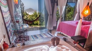 A person relaxing in a bathtub filled with bubbles, with a view of a beach and a pink bed in the background. - Pousada Villa Buena Vista (Imbituba)