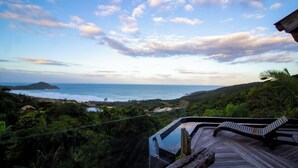 A wooden deck with a glass balustrade overlooking a coastal landscape with a beach and hills. - Pousada Villa Buena Vista (Imbituba)