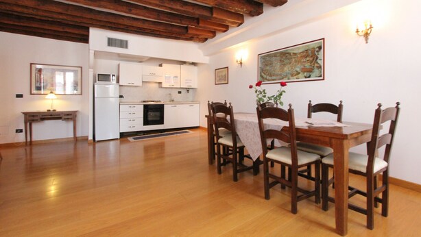 A dining area with wooden chairs and a table, a kitchen in the background, and a painting on the wall. - Apartment Moro 5 (80mt. from Rialto´s Bridge) (Venecia)