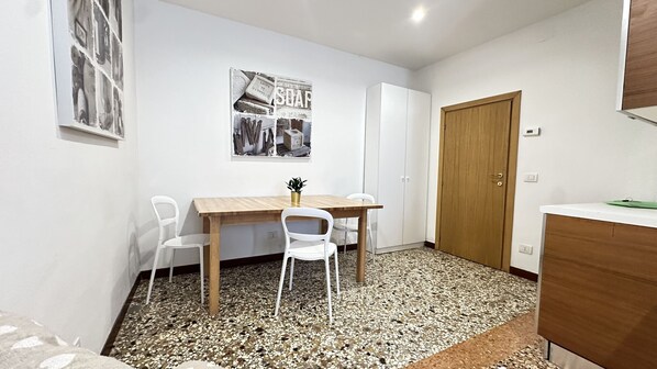 A small room with a wooden table, two white chairs, a potted plant, a door, and a wall-mounted poster.