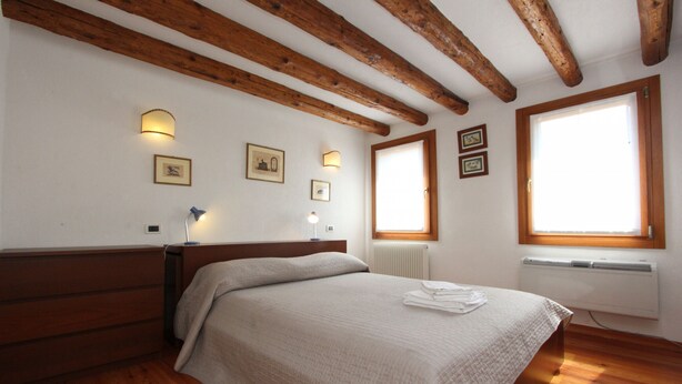 A bedroom with a wooden ceiling, a bed with a white bedspread, a nightstand with a lamp, and framed pictures on the wall.