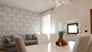 Un coin repas avec une table blanche, quatre chaises et un saladier de fruits. Un canapé gris et un papier peint à motifs se trouvent à l’arrière-plan.