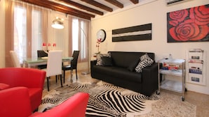 Ein Wohnzimmer mit einer schwarzen Couch, roten Sesseln, einem Zebra-Teppich, einem Esstisch mit Stühlen, einer Wanduhr und einem Gemälde einer roten Rose.