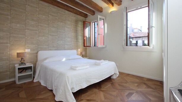 Une chambre avec un plancher en bois, un lit blanc, une table de chevet avec une lampe et une fenêtre donnant sur des immeubles.