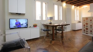 Un vaste espace de vie doté d’un plancher en bois, d’une table à manger et de chaises. Un téléviseur est fixé au mur, et une fenêtre offre une vue sur l’extérieur.
