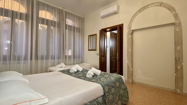 A hotel room with two beds, a large window with sheer curtains, an air conditioning unit, and a door leading to another room. - Apartment Canova Canal View (Venecia)
