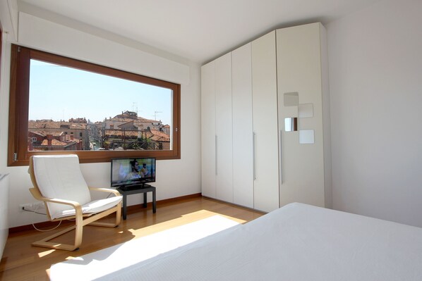 Quarto com uma janela grande, uma cama, uma televisão sobre uma mesinha e roupeiros brancos.