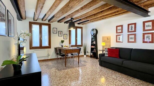 A living room with a black sofa, wooden beams, a dining area with a table and chairs, and framed pictures on the walls. - San Giovanni (Venice Historical Centre) (Venecia)