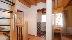 Un escalier en bois mène à une pièce dont le sol est recouvert de carreaux, qui possède une fenêtre avec des rideaux et dont on aperçoit la porte de la salle de bain.