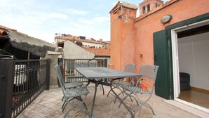Une terrasse extérieure avec une table et des chaises, un portail en métal et un bâtiment aux murs rouges surmonté d’une tour.