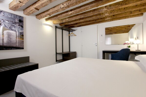 A hotel room with a large bed, a desk with a chair, a wooden ceiling, a wall-mounted mirror, and a painting.