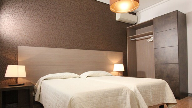 Ein Hotelzimmer mit zwei Betten, einem Nachttisch mit Lampe, einem Kleiderschrank und einer Wand mit Muster.