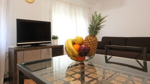 Ein Wohnzimmer mit einem Couchtisch aus Glas, einem Flachbildfernseher auf einem Holzständer, einem braunen Sofa und einer Obstschale auf dem Tisch.
