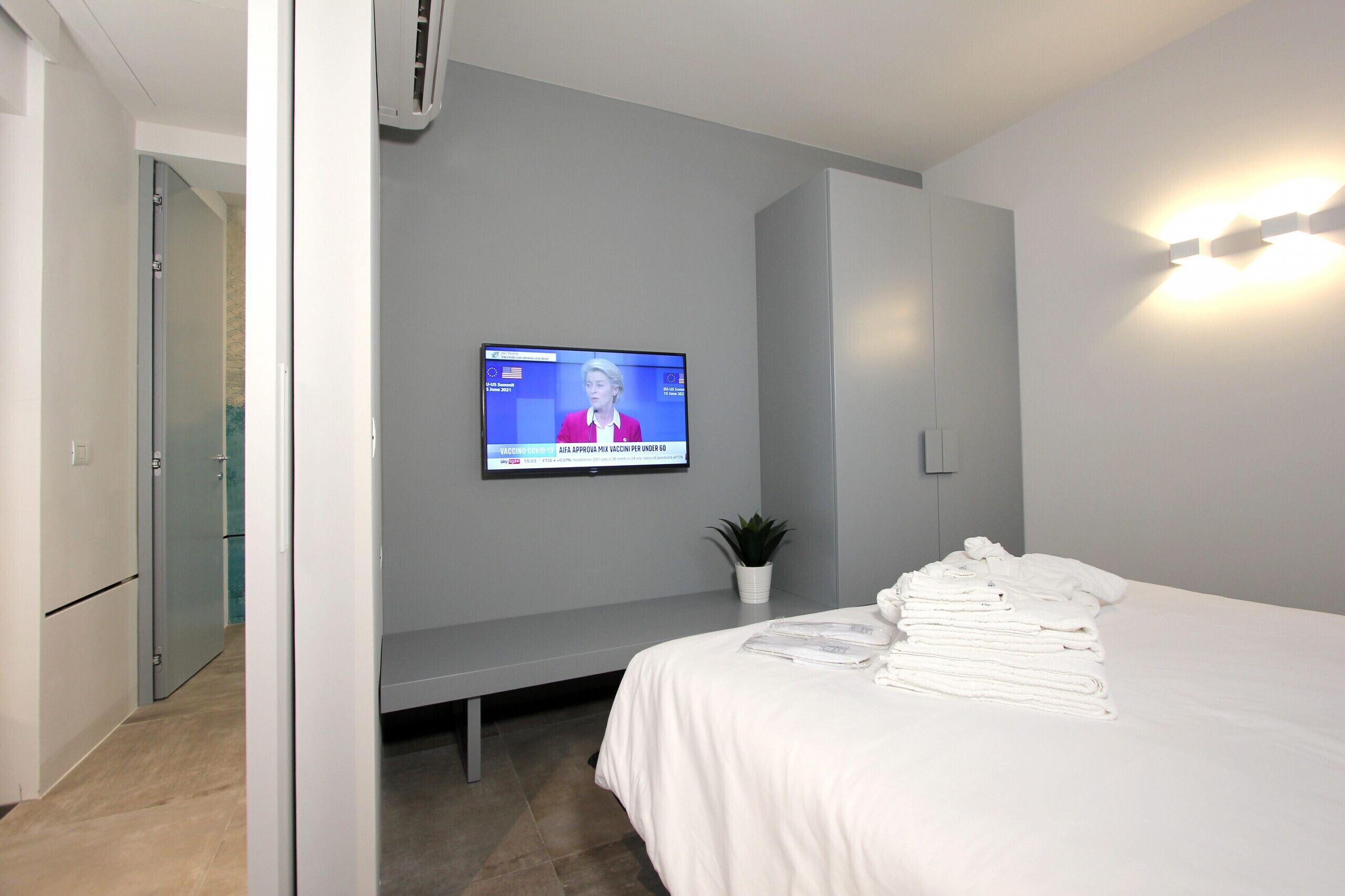 A hotel room with a bed, a TV mounted on the wall, a small potted plant, and a bench.