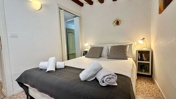 A neatly made bed with white and grey linens, a bedside table with a lamp, and a bookshelf with books.