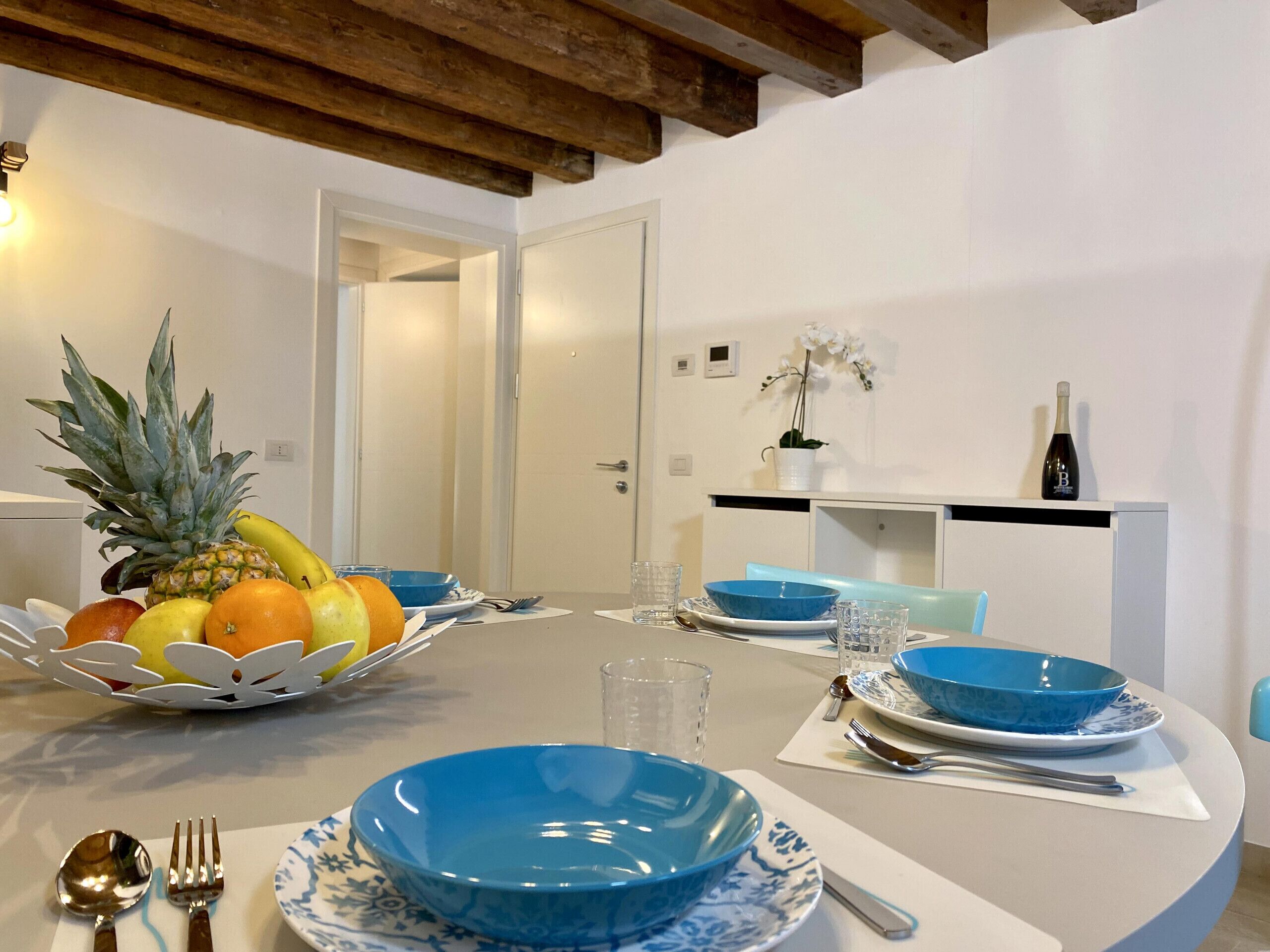 A dining area with a white table set for two, featuring blue and white plates, bowls, and cutlery. A fruit basket with pineapple, banana, and orange is on the table.