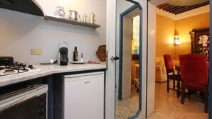 Uma cozinha compacta com fogão, cafeteira e uma pequena geladeira. É possível ver uma área de jantar com mesa e cadeiras através de uma porta aberta.