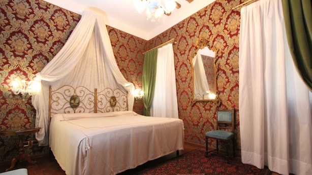 Quarto com cama de dossel, piso de madeira, carpete estampado, mesa de cabeceira e uma cadeira.