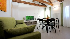 Una sala de estar con sofá verde, vigas de madera, una mesa de comedor con sillas y un televisor sobre un mueble.