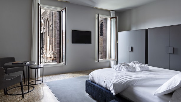 A modern hotel room with a large bed, a desk, and two windows offering views of a brick building.