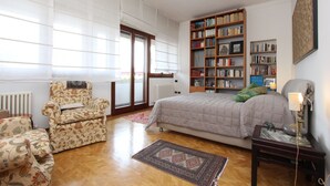 Een slaapkamer met een groot bed, een boekenkast, een stoel en een raam met jaloezieën.