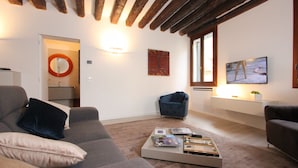 A modern living room with a grey sofa, a coffee table with books and remote controls, a blue armchair, and a wall-mounted TV. - San Polo Canal View, 500m from Rialto´s Bridge (Venecia)