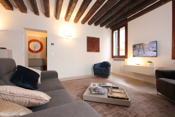 Ein modernes Wohnzimmer mit einem grauen Sofa, einem Couchtisch mit Büchern und Fernbedienungen, einem blauen Sessel und einem an der Wand montierten Fernseher.
