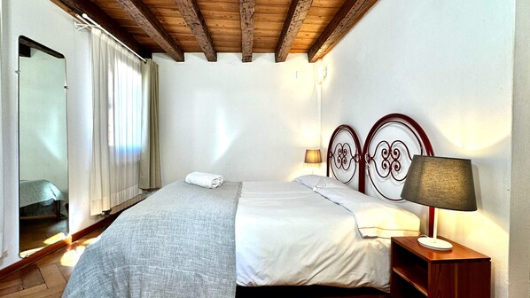 A bedroom with a bed, a nightstand with a lamp, a mirror, and a window with curtains. - Miracoli Terrace (300mt from Rialto´s Bridge) (Venecia)
